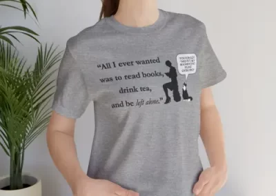 A woman wearing a t-shirt with the quote "All I ever wanted was to read books, drink tea, and be left alone." as well as a picture of characters Lily and Sir Kipling. Sir Kipling has a speech bubble that says "You forgot 'And pet my magnificent feline overlord'".